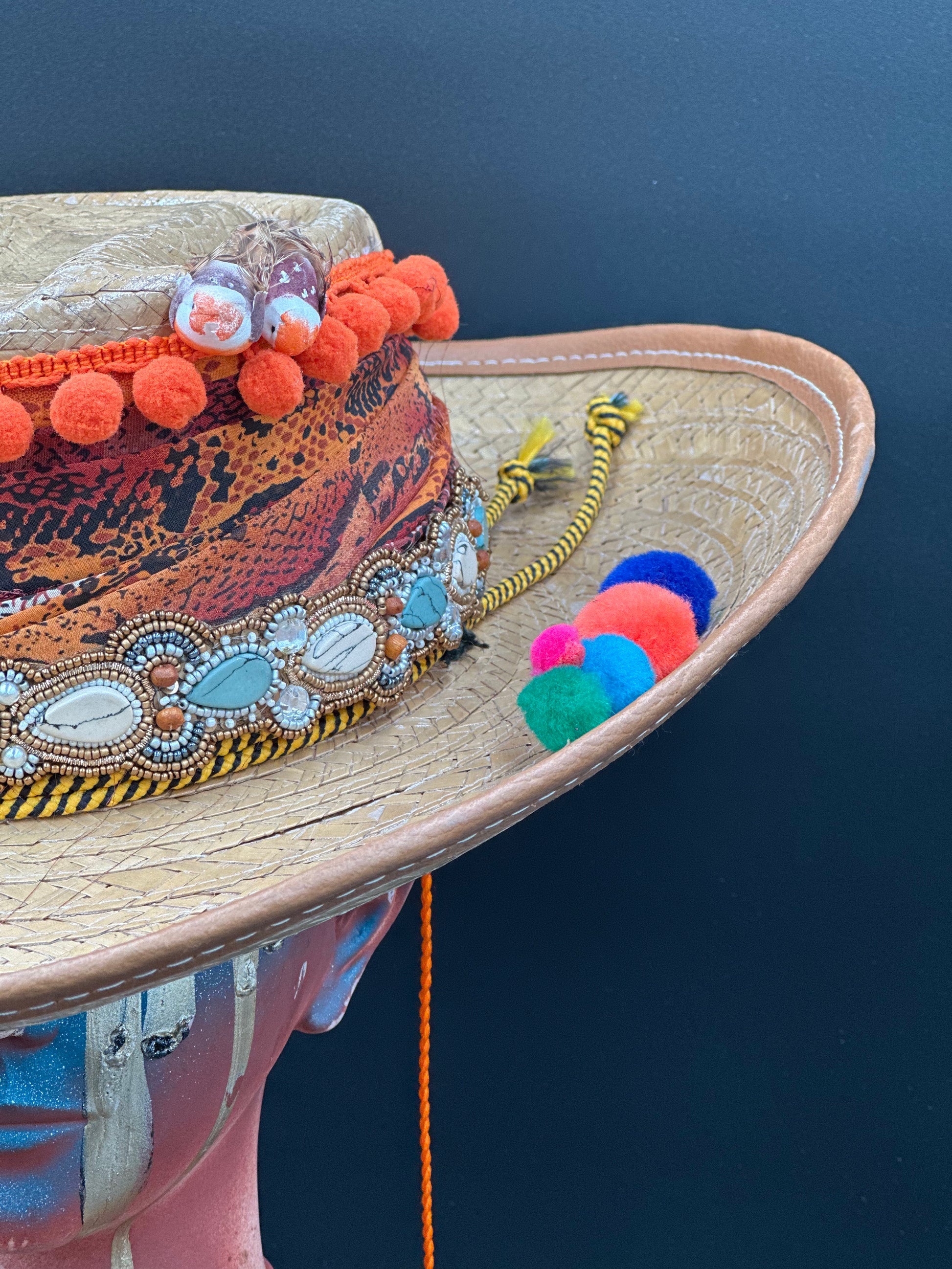 Colombia Straw Hat with colorful embellishments, including orange pom-poms, beads, and a bird design, on a mannequin head. Front view showing the full brim and crown.