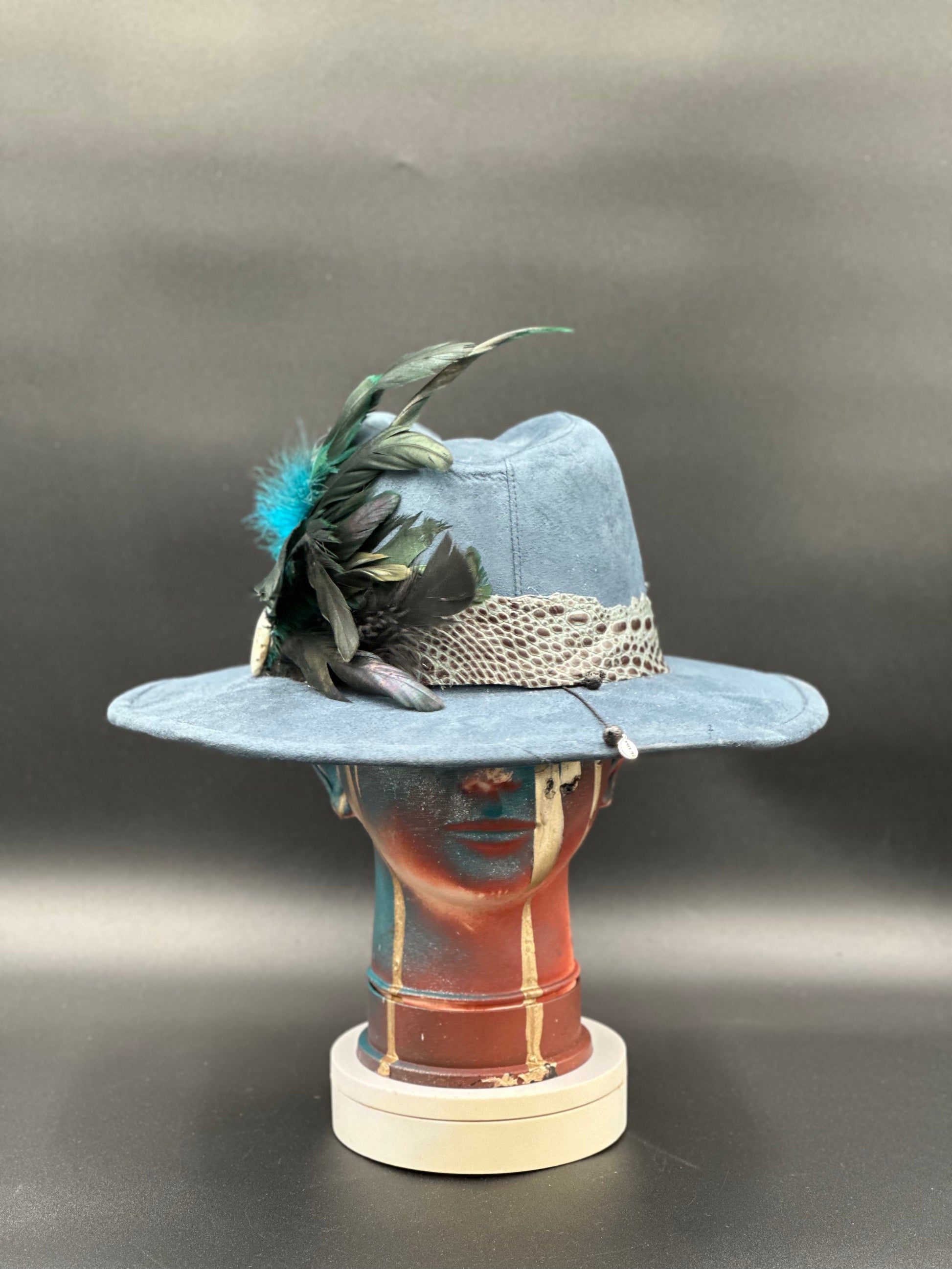 Lisboa Blue Suede Fedora Hat with a feather bouquet, crocodile leather wrap, and a stone embellishment displayed on a mannequin head. Front view showing the hat's full design and shape.